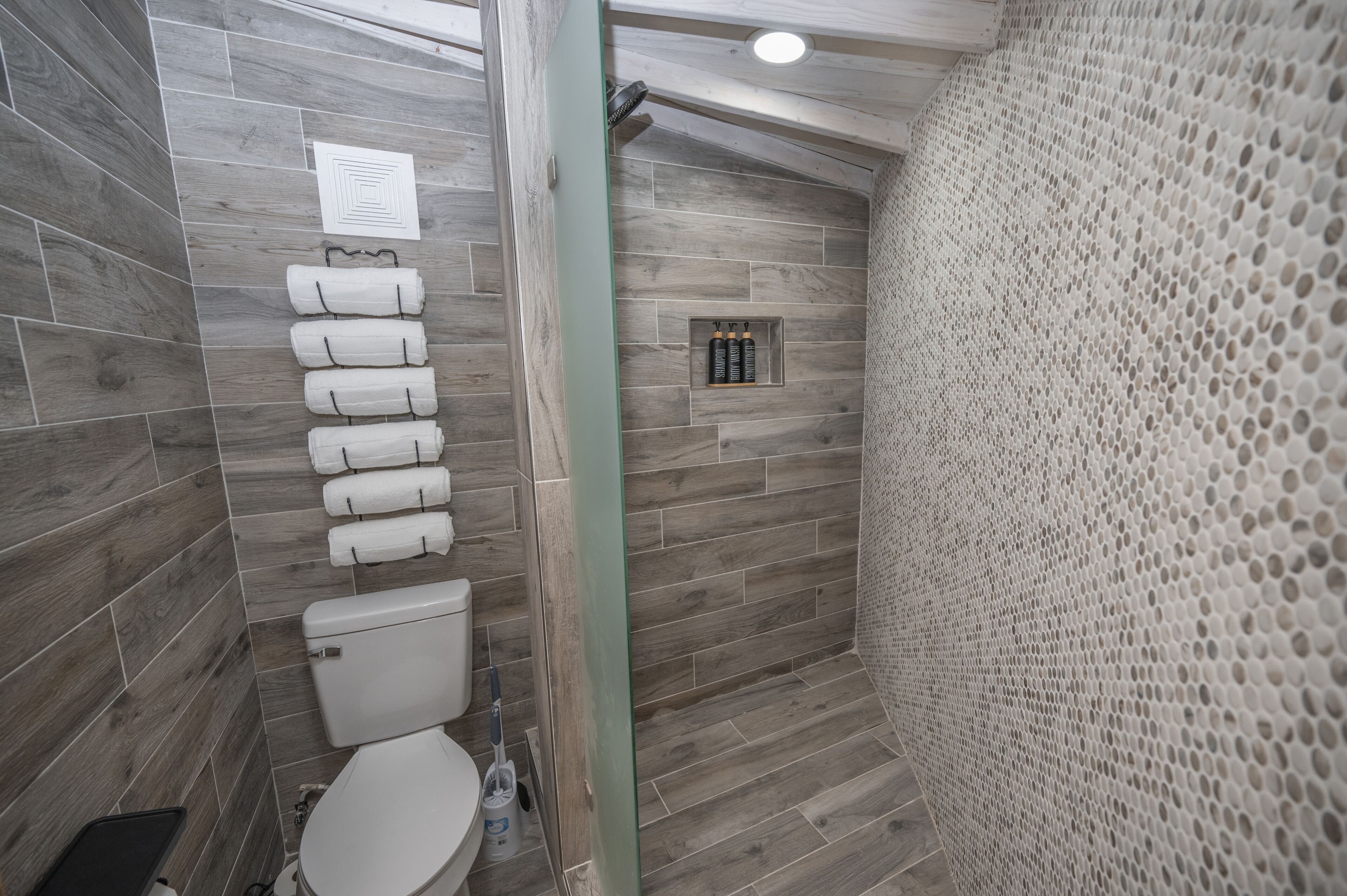 A bathroom with wooden flooring, a toilet, and a shower area with a perforated wall.