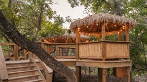 Una casa en el árbol de madera con techo de paja, construida alrededor de un gran árbol. Tiene una escalera de madera y una plataforma con barandilla.