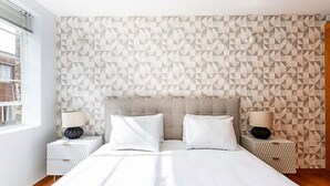 Une chambre d’hôtel moderne avec un mur orné d’un motif géométrique, un grand lit avec une literie blanche, deux tables de chevet avec des lampes et une fenêtre laissant entrer la lumière naturelle.