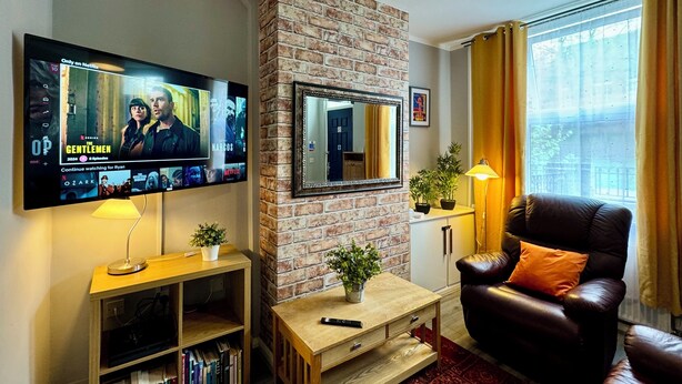 A living room with a brick wall, a flat-screen TV mounted on the wall, a leather sofa, a wooden coffee table, and a bookshelf. - Waterloo House (Nottingham)