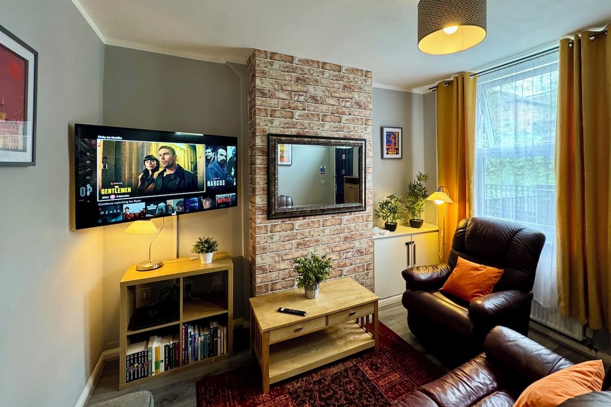 A living room with a brick accent wall, a flat-screen TV, a leather sofa, a coffee table, and a bookshelf.