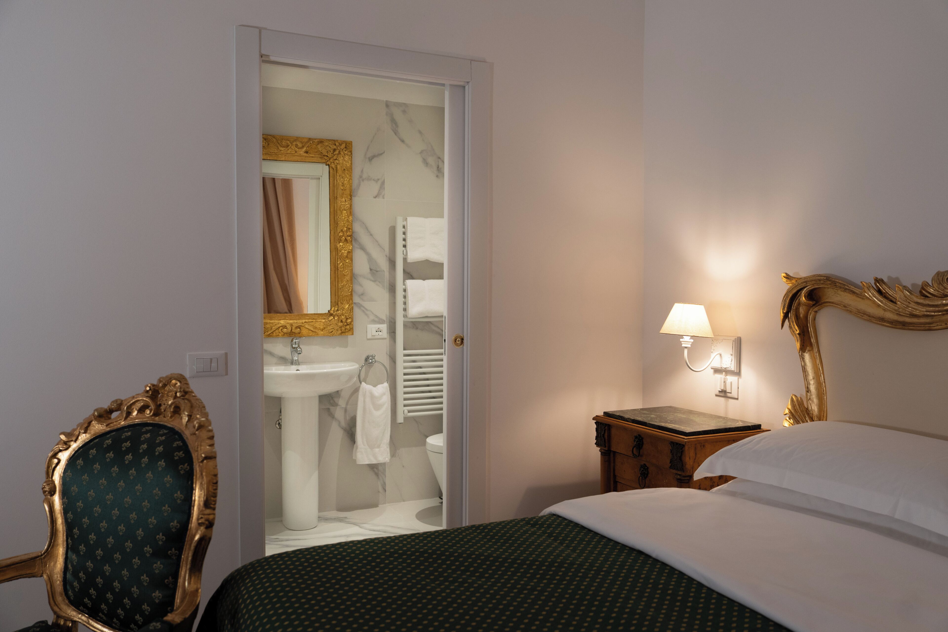 A hotel room with a bed, a nightstand with a lamp, a chair, a bathroom visible through an open door, and a marble wall.