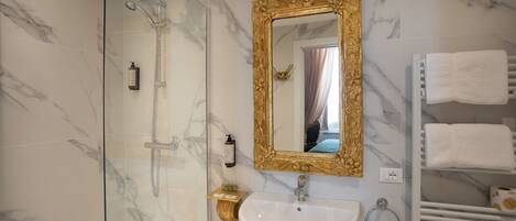 Een badkamer met een douche met glazen wand, een marmeren wand, een spiegel met gouden lijst, een witte wastafel en een handdoekrek met meerdere handdoeken.