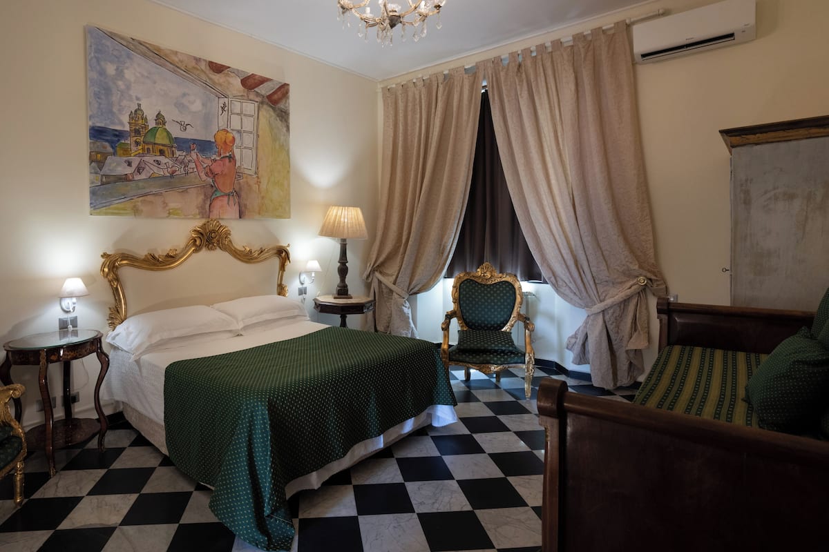 A hotel room with a checkered floor, a bed with a green bedspread, a painting on the wall, a chandelier, and a window with curtains.