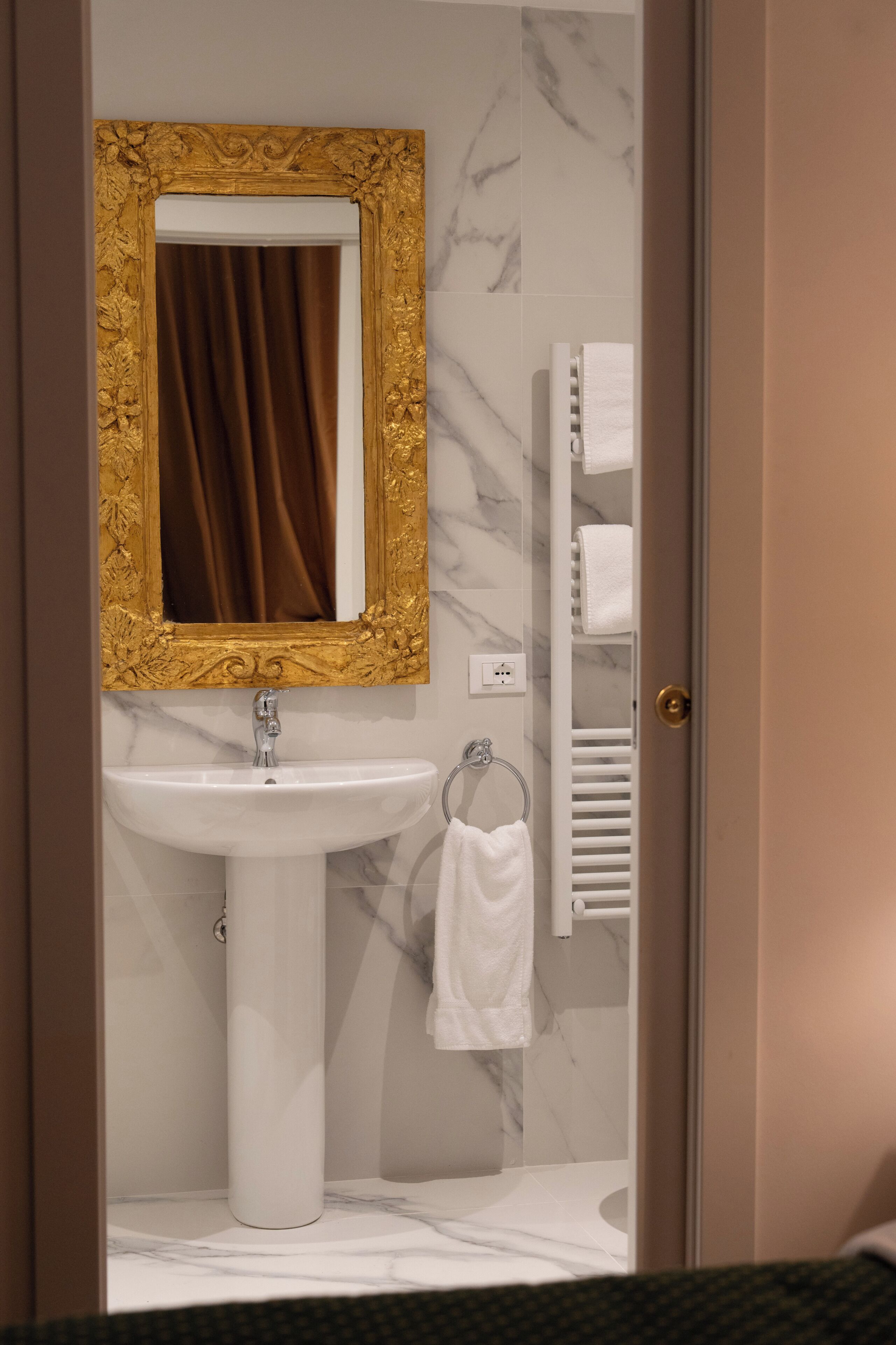Un bagno con un lavandino bianco, uno specchio con cornice dorata, un portascopevoli e un asciugamano bianco.