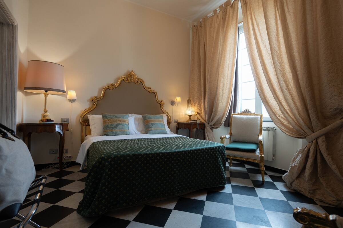 A hotel room with a checkered floor, a bed with a green bedspread, a gold-framed headboard, and a chair with a green cushion.