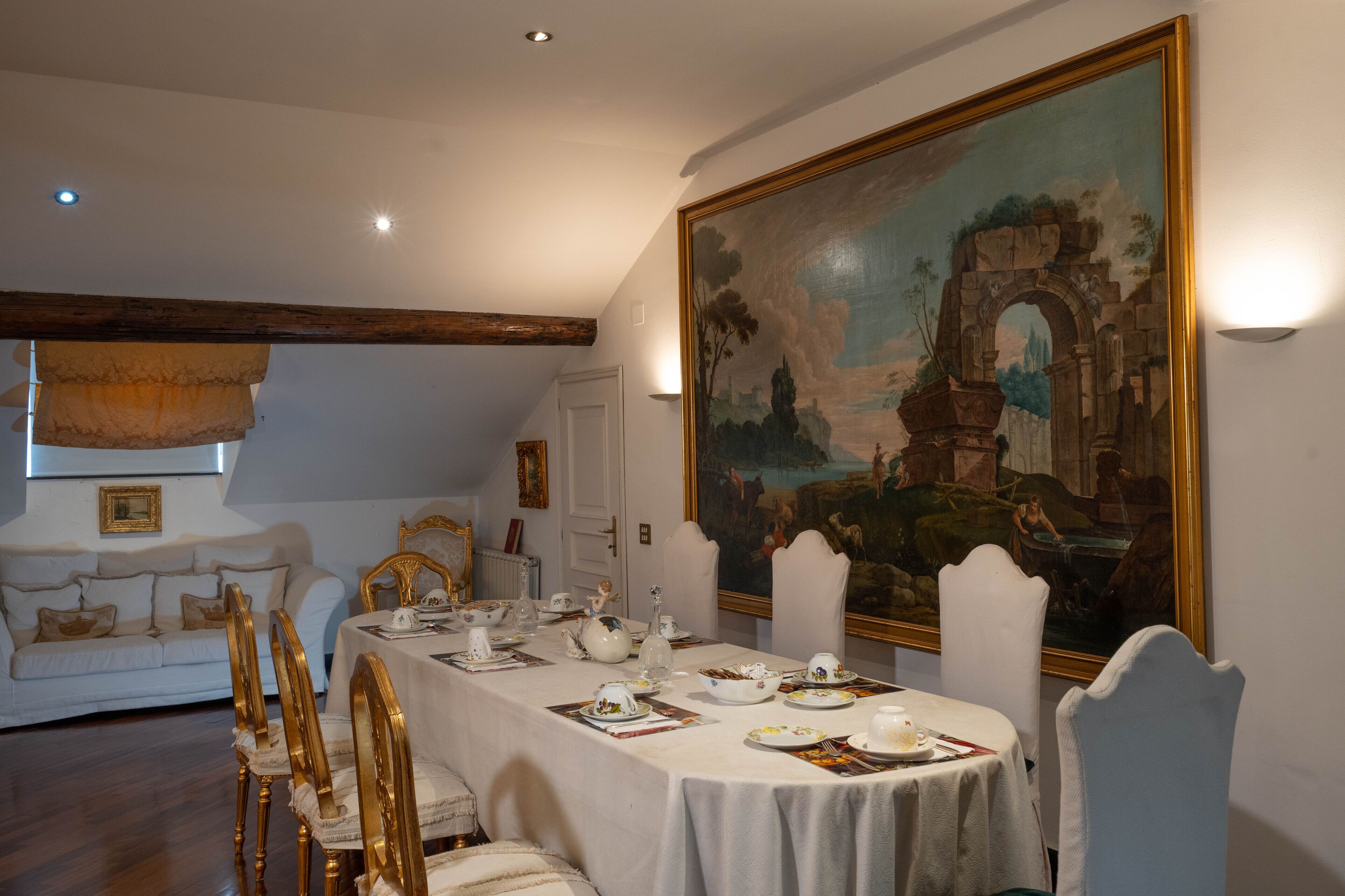 Ein Esszimmer mit einem langen Tisch, der für eine Mahlzeit gedeckt ist, einer weißen Couch im Hintergrund und einem großen Landschaftsbild an der Wand.