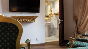 Una habitación con un televisor montado en la pared, un espejo con marco dorado, un baño con lavamanos de mármol y una puerta que da a otra habitación.