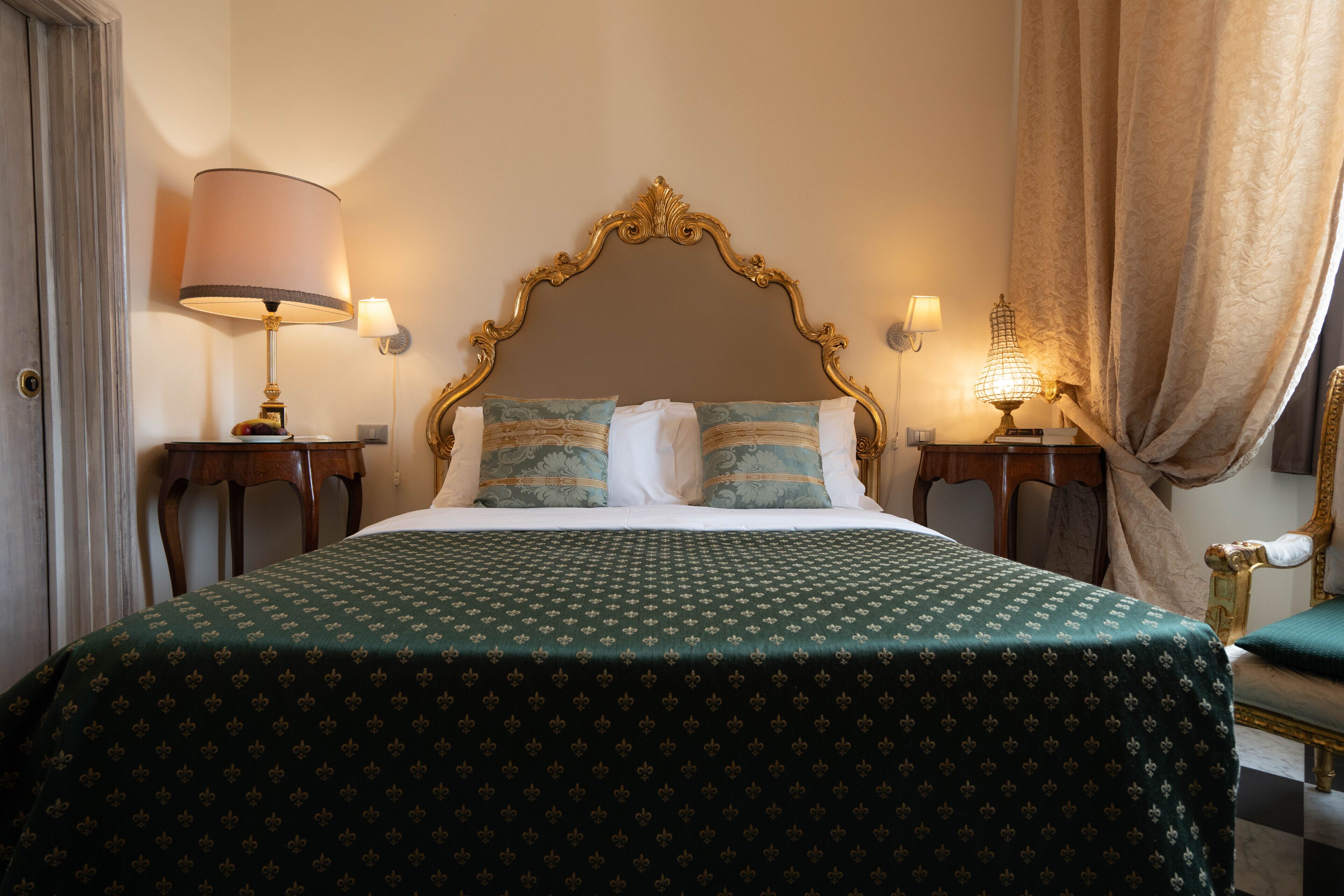 Una cama bien hecha con una colcha verde estampada, dos almohadas y un cabecero con marco dorado. Hay dos mesitas de noche con lámparas, una a cada lado de la cama.