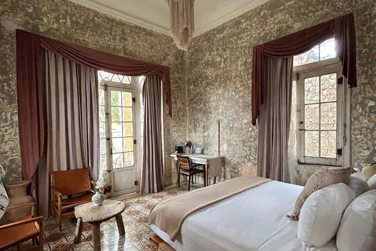 A bedroom with a large bed, a desk, and a chair. There are curtains on the windows, and the walls have a textured pattern.