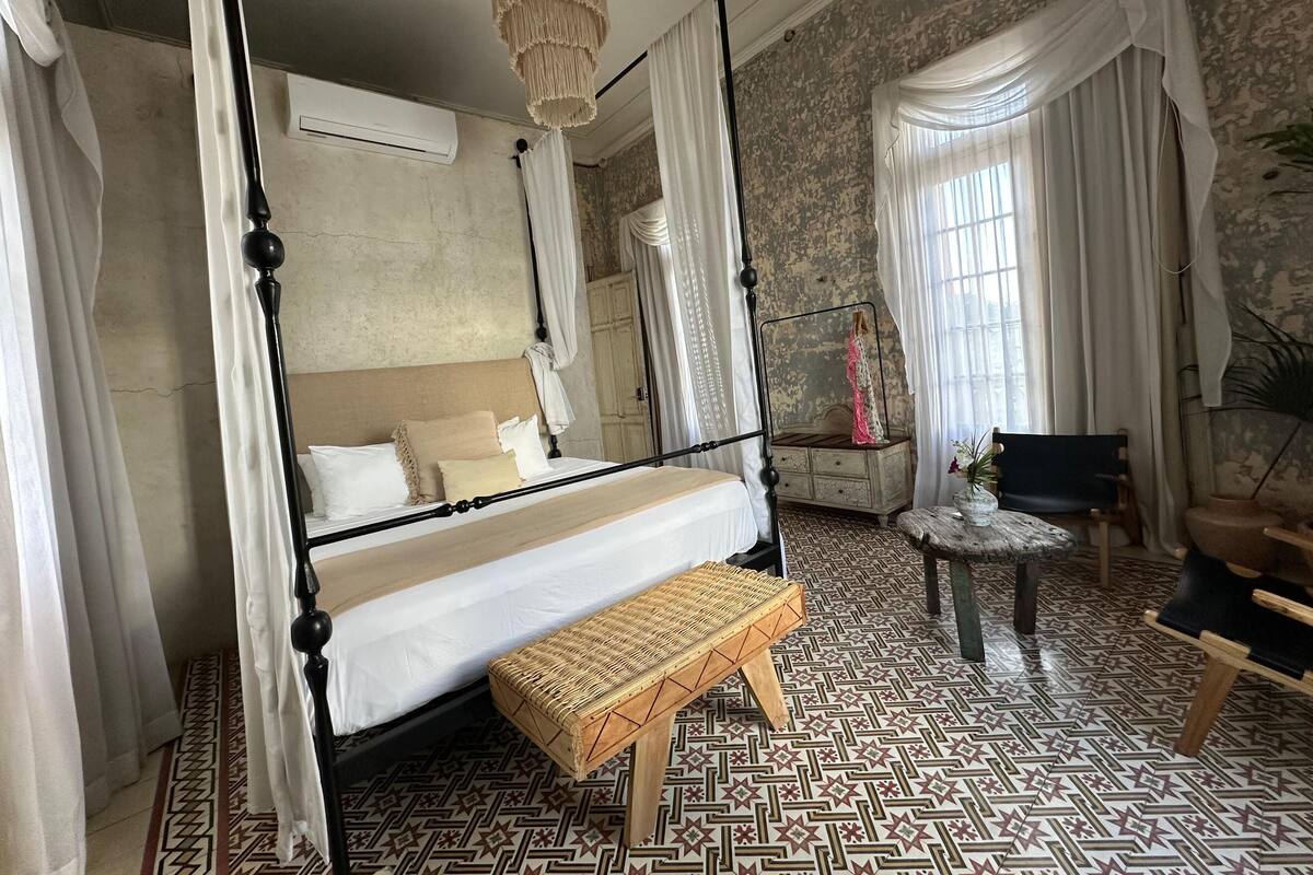 A four-poster bed with a canopy, a small round table, a bench, and a chair in a room with patterned flooring and a stone wall.