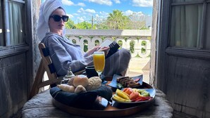 A person sitting on a wooden chair with a tray of food and a drink on a balcony. - Historic House Hotel Boutique (Mérida)