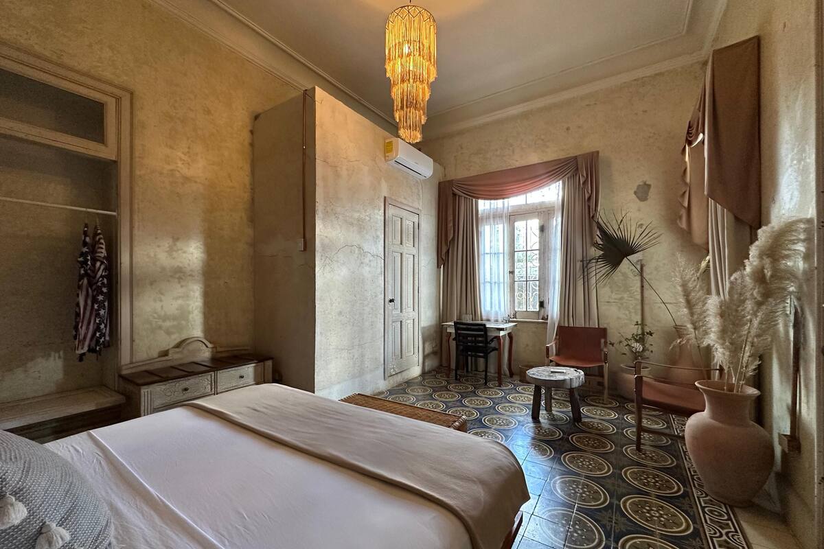 A bedroom with a bed, a desk, and a chair. There is a window with curtains, a hanging light fixture, and a potted plant.