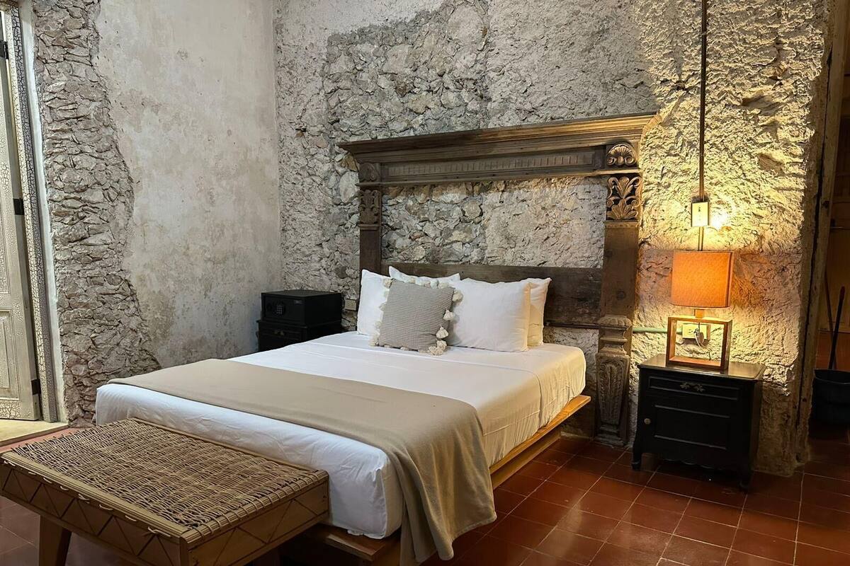 A bedroom with a stone wall, a wooden bed frame, a bedside table with a lamp, and a woven bench.