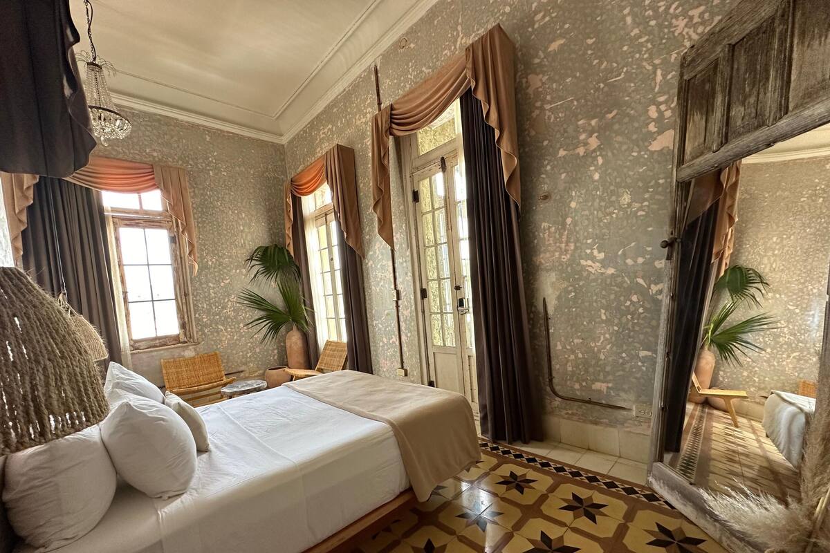 A room with a bed, two chairs, a large mirror, and a potted plant.