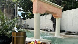 A poolside setting with a wooden table holding a fruit platter and a metal bucket, likely for ice or water, near a fountain. - Historic House Hotel Boutique (Mérida)
