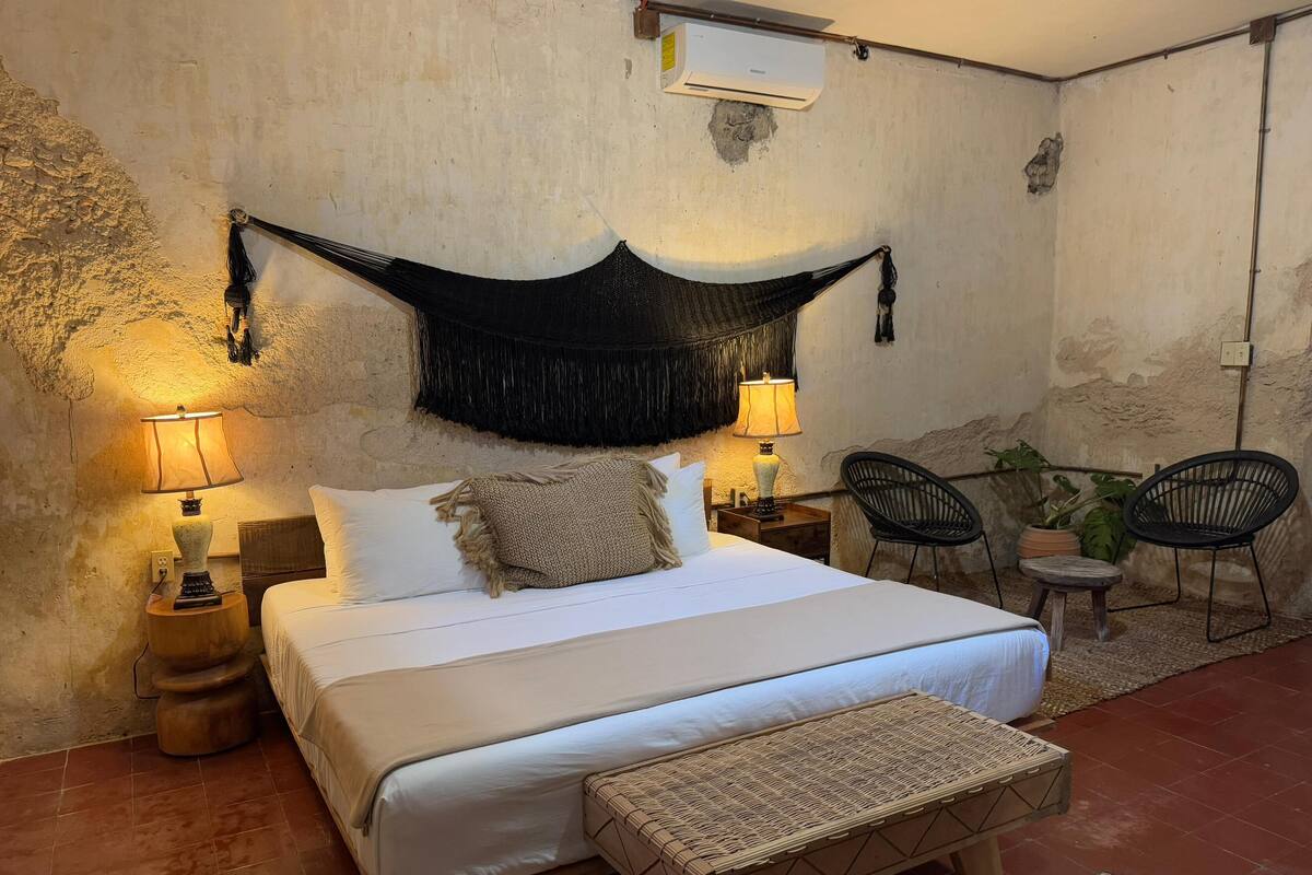 A bedroom with a bed, two bedside lamps, a hanging black tapestry, a small wooden stool, and two chairs.