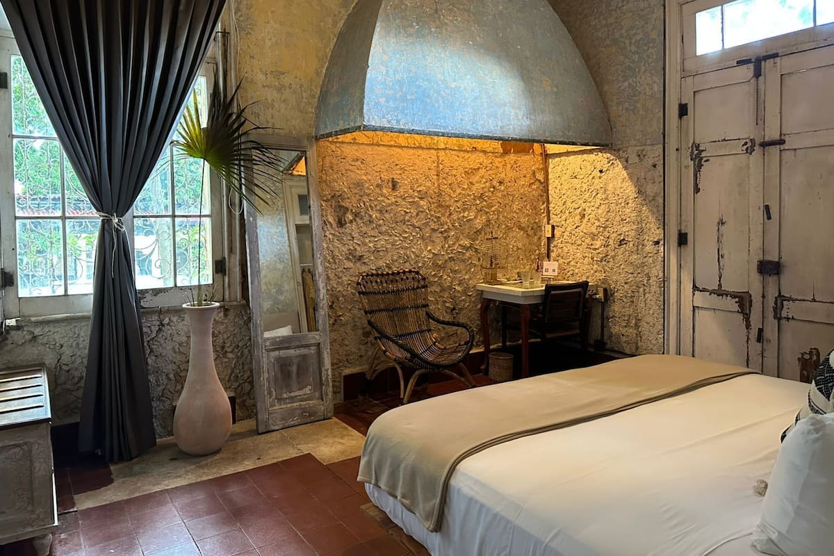 A rustic room with a bed, a table with chairs, a large mirror, and a window with curtains.