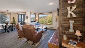 Un salon spacieux avec un canapé en cuir marron, une table basse en bois et une grande fenêtre donnant sur un paysage de arbres et de montagnes.