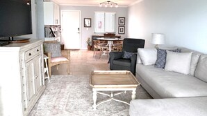 Ein Wohnzimmer mit einer Couch, einem Couchtisch und einem Fernseher. Im Hintergrund befindet sich ein Essbereich mit Tisch und Stühlen.