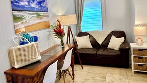 A living room with a wooden table, white chair, brown leather sofa, and a lamp. - Adventurers' Dream Launchpad: near Shops & Beaches: SUPs, Bikes, Snorkel & More! (Princeville)