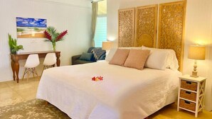 A neatly arranged bedroom with a large bed, a desk with chairs, and a decorative wall panel. - Adventurers' Dream Launchpad: near Shops & Beaches: SUPs, Bikes, Snorkel & More! (Princeville)