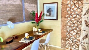 A dining area with a wooden table, two chairs, a vase with flowers, and a framed picture on the wall. - Adventurers' Dream Launchpad: near Shops & Beaches: SUPs, Bikes, Snorkel & More! (Princeville)