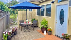 A patio with a table and chairs under a blue umbrella, surrounded by potted plants and a wooden fence. - Adventurers' Dream Launchpad: near Shops & Beaches: SUPs, Bikes, Snorkel & More! (Princeville)