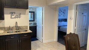 A modern kitchen with dark cabinets, a granite countertop, and a sink. There is a dining area with a brown chair and a small table. A bedroom is visible through an open door, featuring a bed and a nightstand. - Private Two Bedroom, Two TVs, One Bathroom Casita! (Las Vegas)