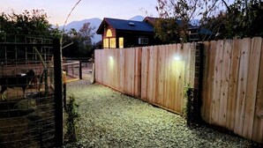 A fenced outdoor area with a gravel path leading to a wooden gate, a wooden fence, and a house with a dark roof and lit windows.