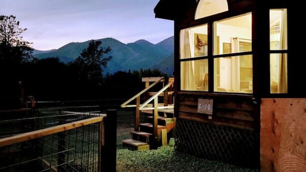 A wooden cabin with a staircase leading to a windowed entrance, surrounded by a metal fence and a mountainous backdrop.