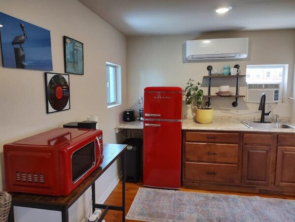 Una cocina compacta con un refrigerador rojo, un microondas y un lavamanos. Hay aires acondicionados instalados en la pared y una ventana que proporciona luz natural.