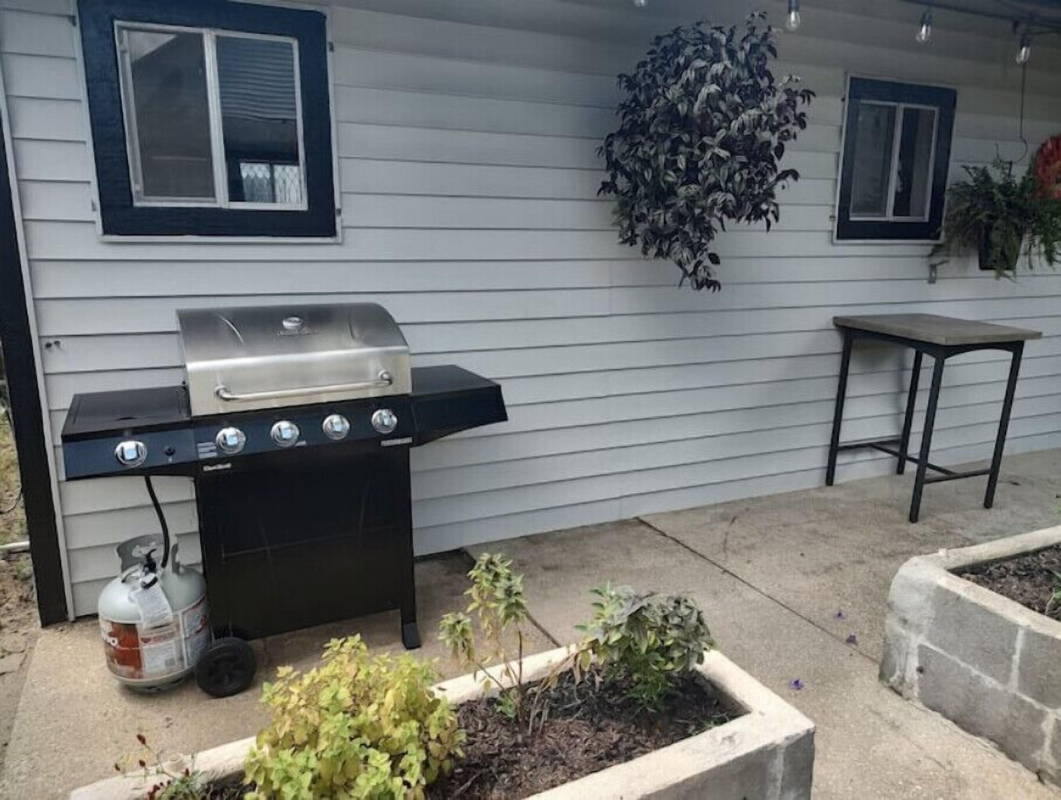A barbecue grill with a gas tank and a table with a chair, positioned in front of a house with white siding and dark window frames.