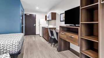 A compact hotel room with a bed, a desk, a chair, a television, and a kitchenette.
