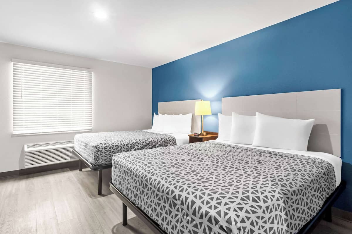 Habitación de hotel con dos camas, una pared azul de acento, una ventana con persianas y una mesita de noche con lámpara.