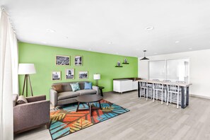 A modern living room with a green accent wall, a sofa, a coffee table, and a dining area with a white table and chairs. - Woodspring Suites South Houston Hobby (Houston)