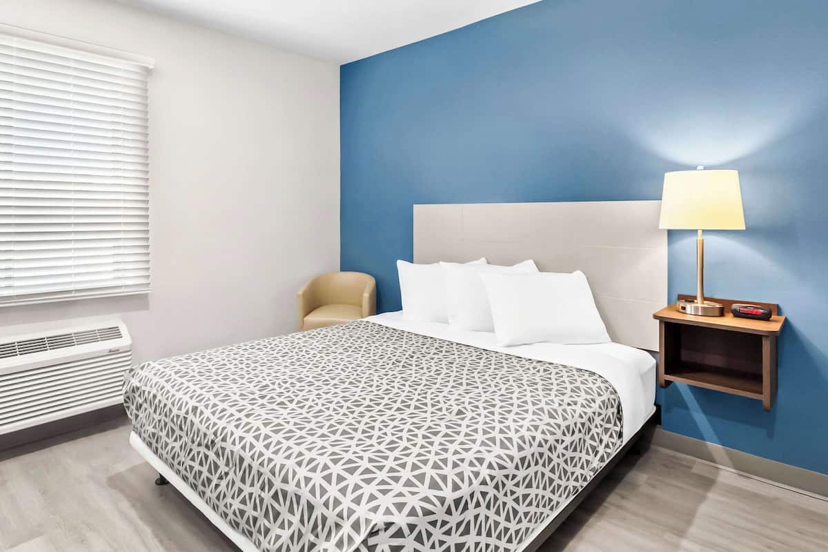Una habitación de hotel con una cama, una mesita de noche con lámpara, una silla y una ventana con persianas.