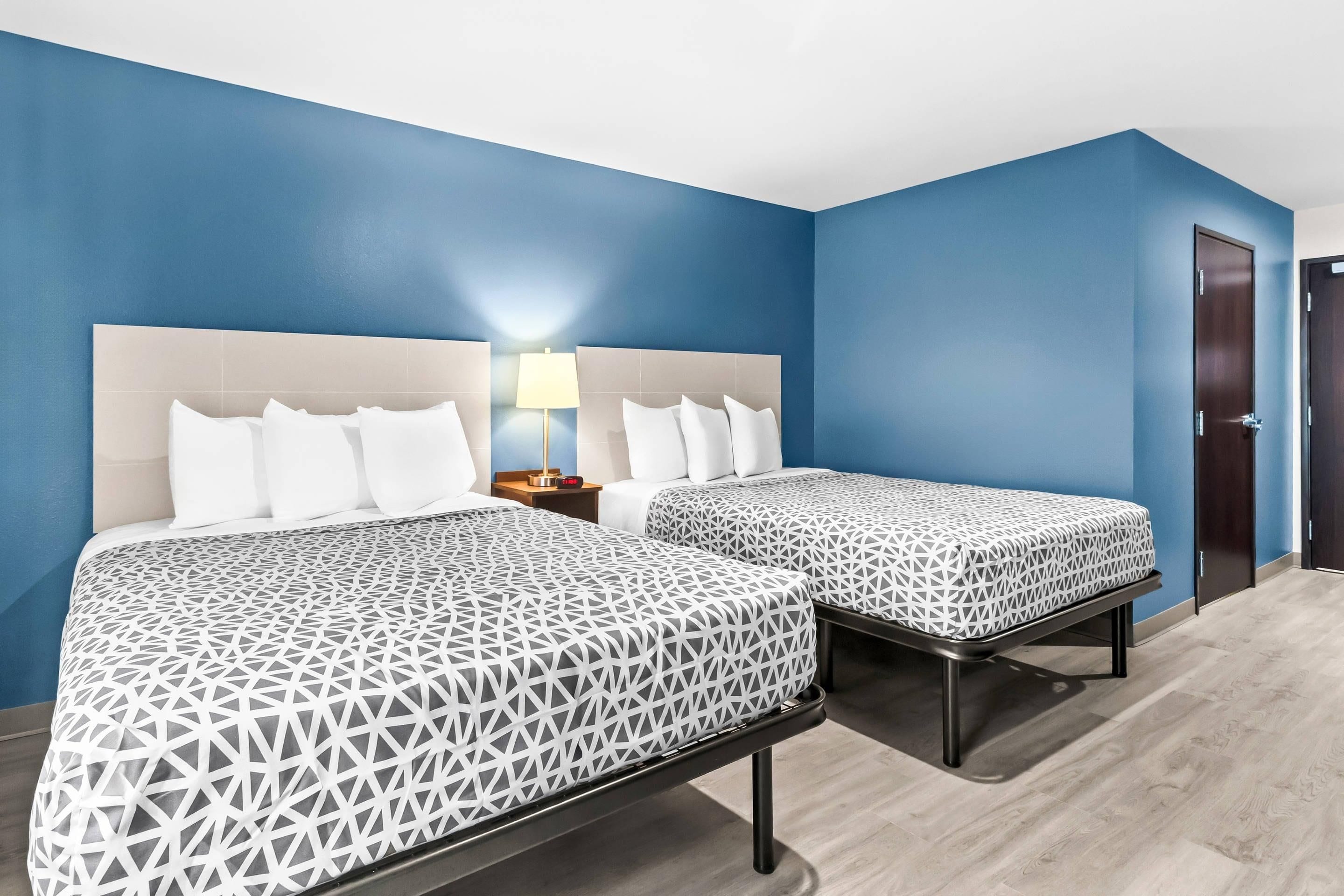 Quarto de hotel com duas camas, uma mesa de cabeceira com abajur e uma porta.