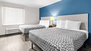 A hotel room with two beds, a blue accent wall, a window with blinds, and a bedside table with a lamp. - Woodspring Suites South Houston Hobby (Houston)