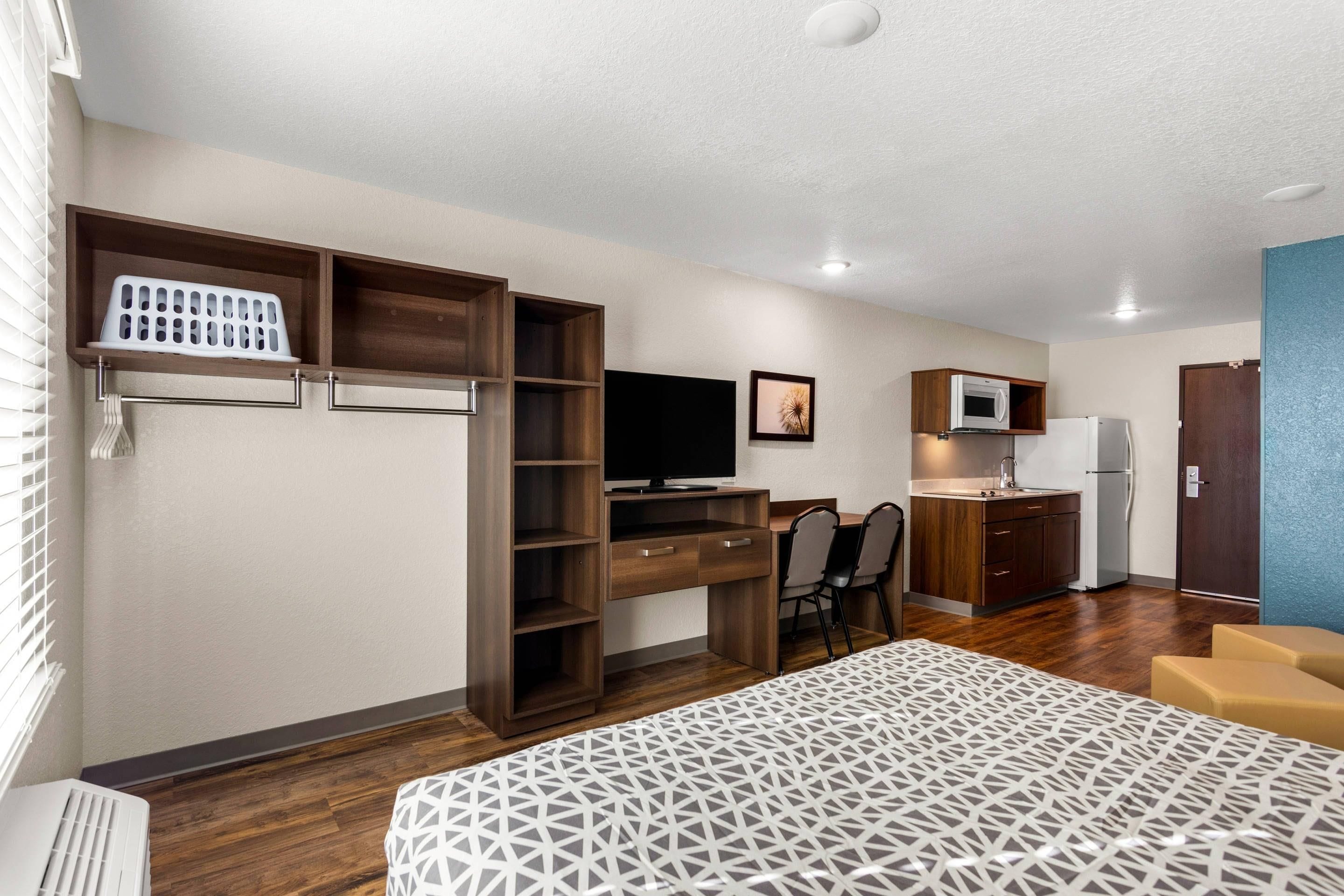 A compact hotel room with a bed, a desk with a computer, a TV, a kitchenette, and a seating area.
