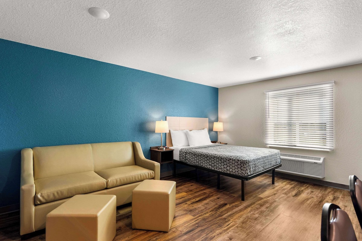 Habitación de hotel con una pared azul de acento, una cama con colcha estampada, un sofá color beige, un pequeño reposapiés, una mesita de noche con lámparas y una ventana con persianas.