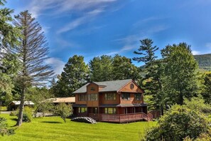 A two-story wooden house with a porch, surrounded by lush greenery and trees.