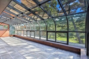 A modern, curved glass structure with a wooden bench and tiled floor, offering a view of a green landscape.