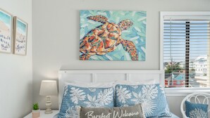 Un dormitorio con una cama que presenta un estampado floral azul y blanco, un cojín con el texto 'Barefoot Welcome', un cuadro colgado en la pared de una tortuga marina y una ventana con persianas.