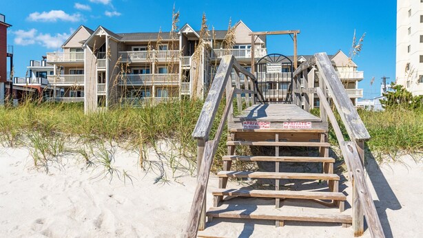 Un paseo marítimo de madera que conduce a un edificio frente a la playa, con varios balcones y un letrero que indica acceso privado.
