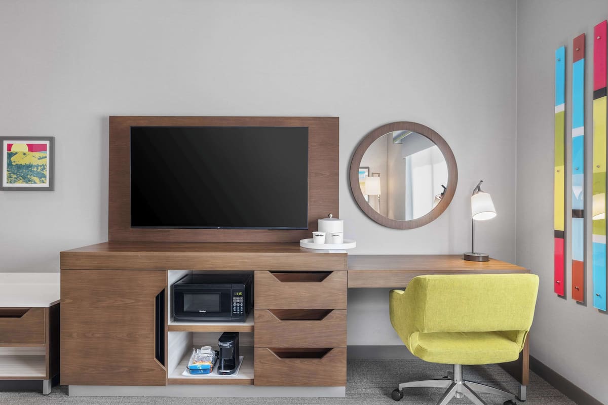 A hotel room with a wooden desk, a flat-screen TV, a microwave, a coffee maker, a yellow chair, a round mirror, and colorful lockers.