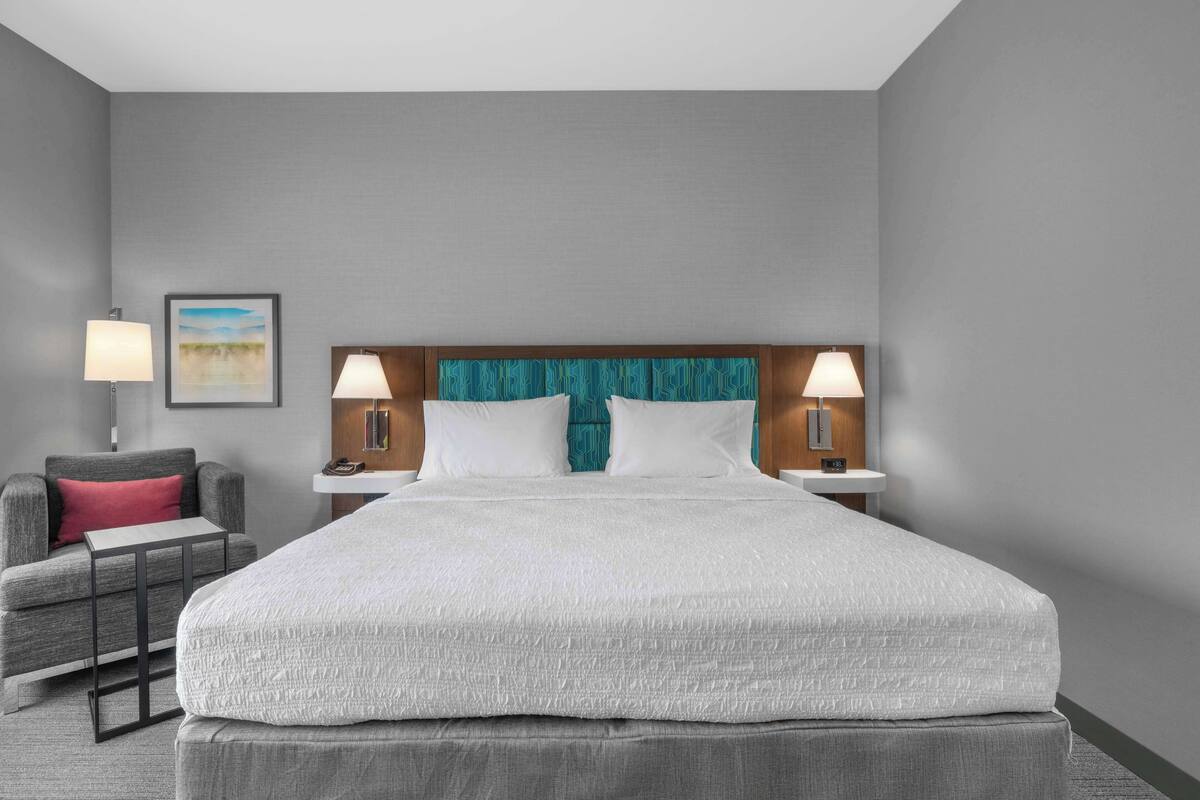 A hotel room with a large bed, two bedside lamps, a small table, a chair, and a framed picture on the wall.