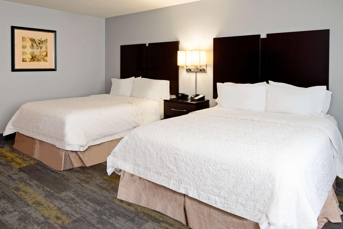 Quarto de hotel com duas camas, uma mesa de cabeceira com abajur e um quadro na parede.