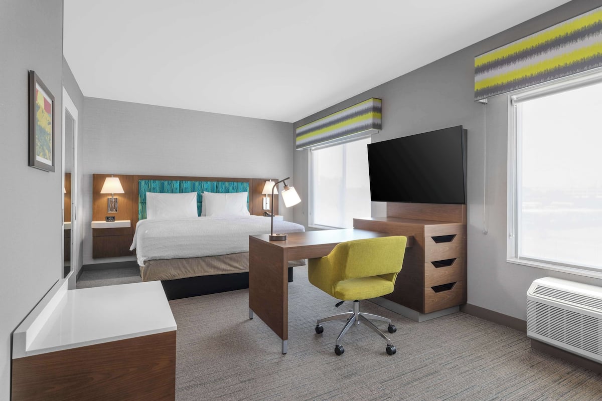 A hotel room with a bed, desk, chair, TV, and large windows.
