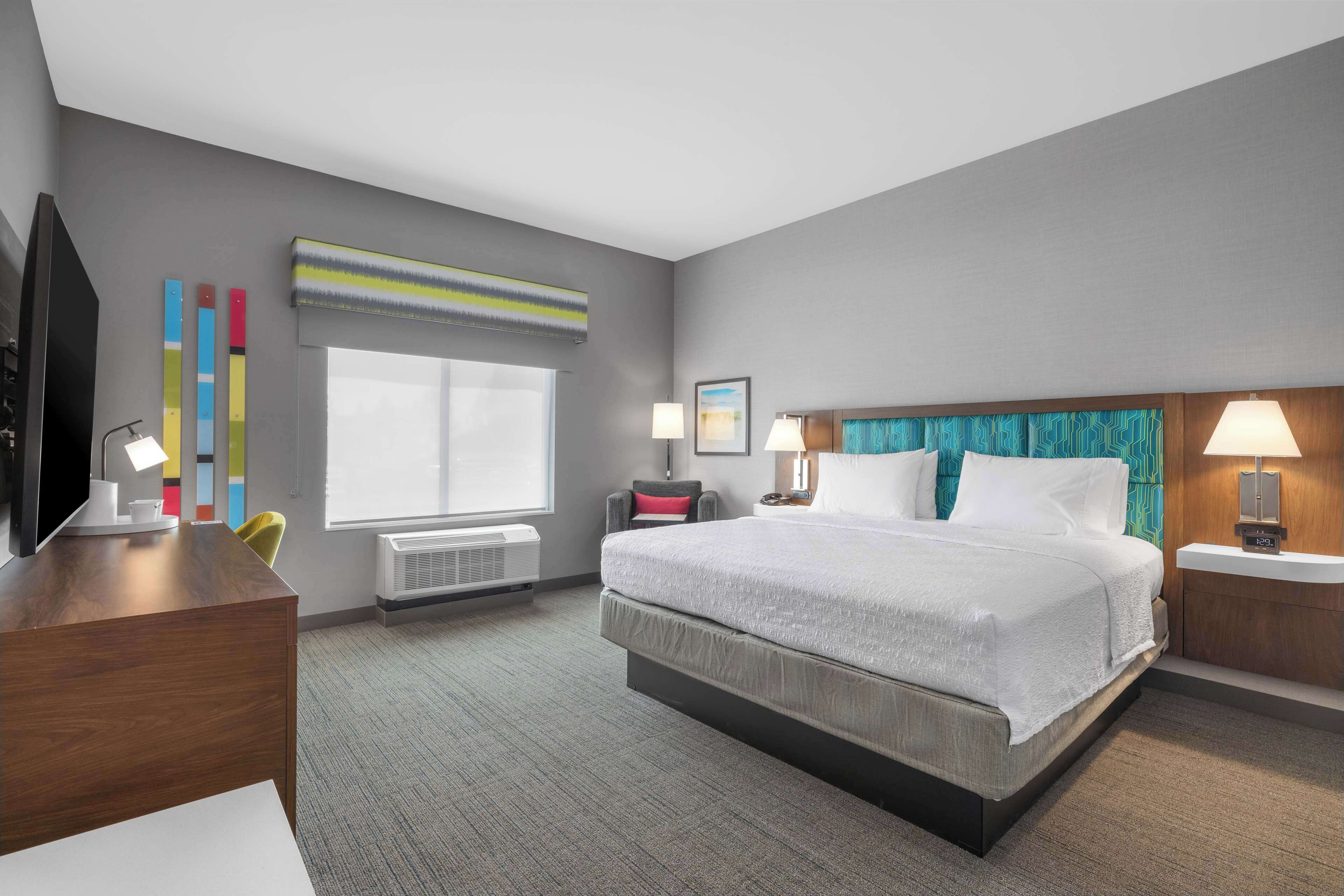 A hotel room with a large bed, a desk with a TV, a window with blinds, and a wall-mounted air conditioner.